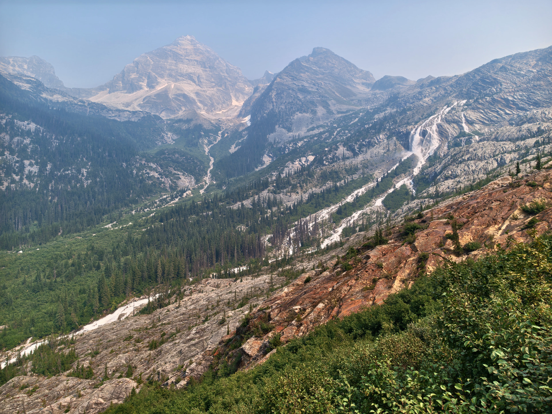 Berglandschaft am Great Glacier Trail mit rötlichem Fels, Wasserfall und durch Waldbrandrauch diesigen Bergen im Hintergrund